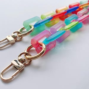 May include: A multi-coloured acrylic chain strap with gold-coloured clasps. The chain links are rectangular and come in various translucent colours, including pink, green, blue, and red. The strap is designed for handbags or other accessories.