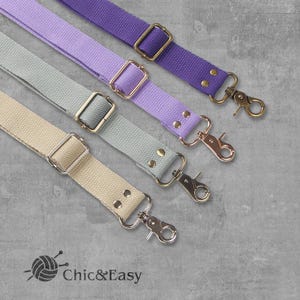 May include: Four different colored canvas straps with gold and silver hardware. The straps are light purple, gray, beige, and dark purple. The straps are all the same width and have a metal clasp on one end and a metal loop on the other end. The straps are arranged in a row from top to bottom, with the light purple strap at the top and the dark purple strap at the bottom.