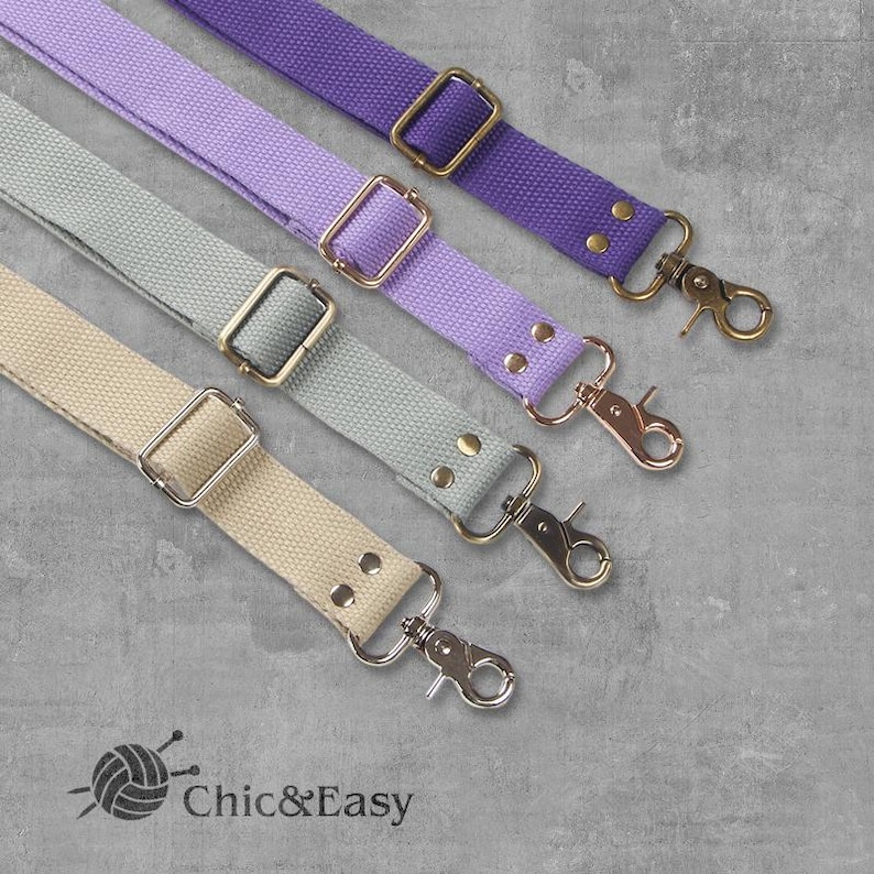 May include: Four different colored canvas straps with gold and silver hardware. The straps are light purple, gray, beige, and dark purple. The straps are all the same width and have a metal clasp on one end and a metal loop on the other end. The straps are arranged in a row from top to bottom, with the light purple strap at the top and the dark purple strap at the bottom.