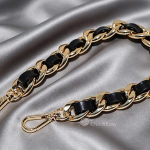Peut inclure: Une bandoulière en chaîne métallique dorée avec des bandes de cuir noir entrelacées. La sangle est courbée et comprend un fermoir mousqueton à chaque extrémité pour la fixation. Le texte Chic&Easy est visible en bas au centre de l'image.