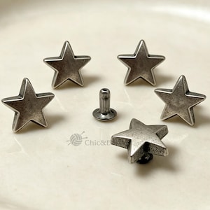 May include: A set of five silver-toned metal star-shaped rivets with a distressed finish. Four of the rivets are positioned with the star face upward, while one central rivet is shown from the side, displaying the cylindrical post and base.