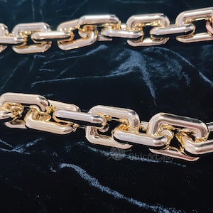 May include: Gold-toned chain links with a polished finish, arranged in two parallel rows against a dark, textured background. The links are rectangular and connected, creating a bold, statement piece. The chain is likely used for jewelry or fashion accessories.