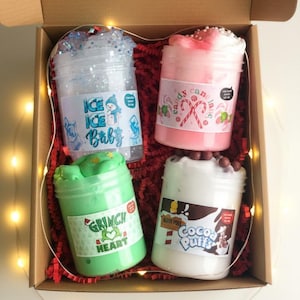 May include: A gift box containing four clear plastic jars filled with colorful slime. The jars have labels with text like "Ice Ice Baby," "Candy Cane Lane," "Grinch Heart," and "Cocoa Puffs." The box is lined with red paper and has fairy lights.