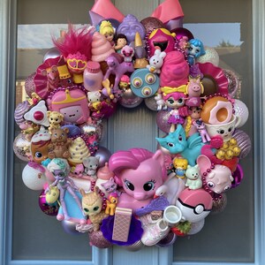 K&ouml;nnte beinhalten: Ein rosa-lilafarbener Weihnachtskranz, der mit verschiedenen kleinen Spielzeugen geschm&uuml;ckt ist, darunter ein rosa Pony, ein blauer Drache und ein rosa-wei&szlig;es Eiswaffelh&ouml;rnchen.
