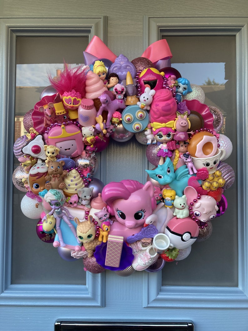 K&ouml;nnte beinhalten: Ein rosa-lilafarbener Weihnachtskranz, der mit verschiedenen kleinen Spielzeugen geschm&uuml;ckt ist, darunter ein rosa Pony, ein blauer Drache und ein rosa-wei&szlig;es Eiswaffelh&ouml;rnchen.