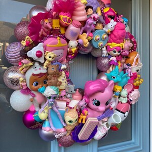 K&ouml;nnte beinhalten: Ein rosa-lilafarbener Weihnachtskranz, der mit verschiedenen kleinen Spielzeugen geschm&uuml;ckt ist, darunter ein rosa Pony, eine Puppe und ein Einhorn. Der Kranz besteht aus glitzernden Kugeln und h&auml;ngt an einer blauen T&uuml;r.