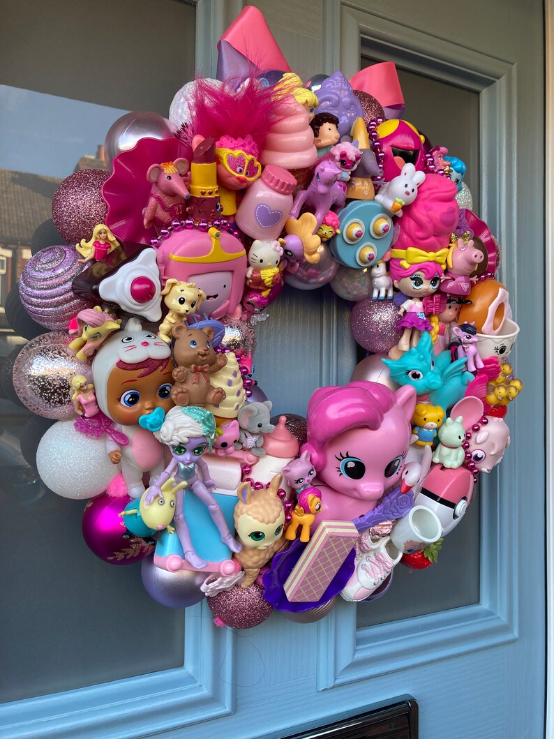 K&ouml;nnte beinhalten: Ein rosa-lilafarbener Weihnachtskranz, der mit verschiedenen kleinen Spielzeugen geschm&uuml;ckt ist, darunter ein rosa Pony, eine Puppe und ein Einhorn. Der Kranz besteht aus glitzernden Kugeln und h&auml;ngt an einer blauen T&uuml;r.