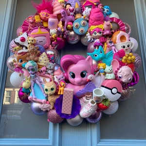 K&ouml;nnte beinhalten: Ein rosa-lilafarbener Kranz, der mit verschiedenen Spielzeugen geschm&uuml;ckt ist, darunter ein rosa Pony, ein Einhorn, eine Puppe und ein Pok&eacute;ball.