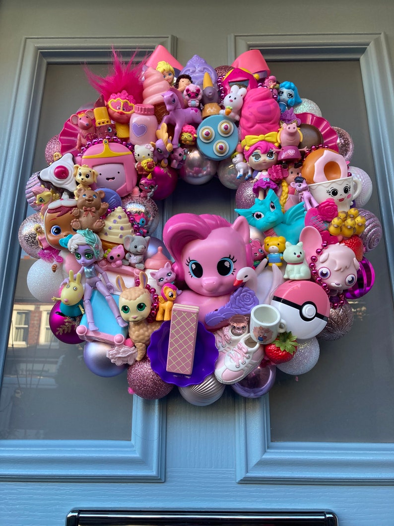 K&ouml;nnte beinhalten: Ein rosa-lilafarbener Kranz, der mit verschiedenen Spielzeugen geschm&uuml;ckt ist, darunter ein rosa Pony, ein Einhorn, eine Puppe und ein Pok&eacute;ball.