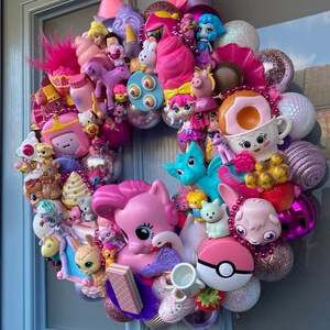 K&ouml;nnte beinhalten: Ein rosa-lilafarbener Kranz, der mit verschiedenen kleinen Spielzeugen geschm&uuml;ckt ist, darunter ein Einhorn, ein Pony, ein Kaninchen, ein Drache, eine Prinzessin und ein Donut. Der Kranz besteht aus verschiedenen Materialien, darunter Plastik, Stoff und Glitzer.