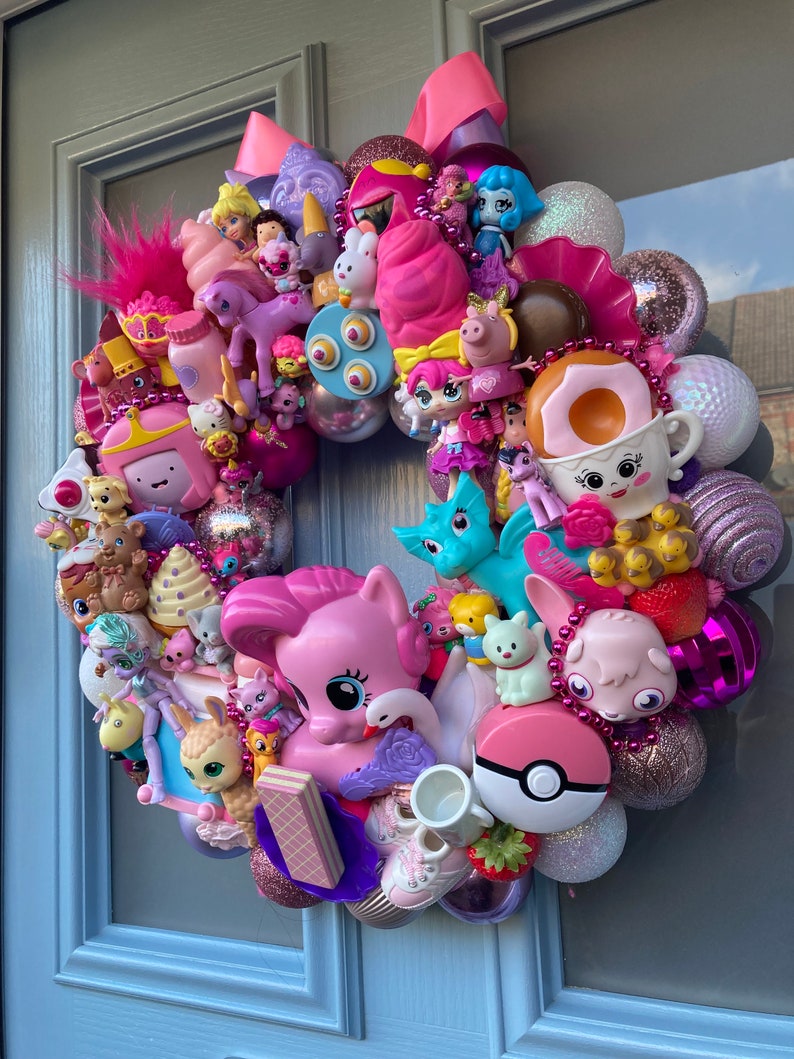 K&ouml;nnte beinhalten: Ein rosa-lilafarbener Kranz, der mit verschiedenen kleinen Spielzeugen geschm&uuml;ckt ist, darunter ein Einhorn, ein Pony, ein Kaninchen, ein Drache, eine Prinzessin und ein Donut. Der Kranz besteht aus verschiedenen Materialien, darunter Plastik, Stoff und Glitzer.