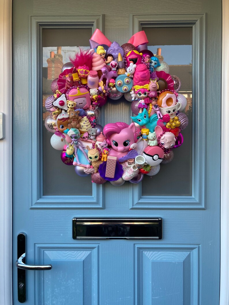 K&ouml;nnte beinhalten: Ein rosa-lilafarbener Kranz, der mit verschiedenen kleinen Spielzeugen geschm&uuml;ckt ist, darunter ein rosa Einhorn, ein rosa Pony, ein blauer Vogel und ein rosa-wei&szlig;er Donut. Der Kranz h&auml;ngt an einer blauen T&uuml;r.