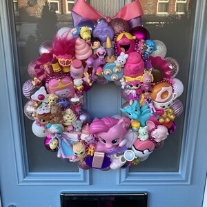 K&ouml;nnte beinhalten: Ein rosa-lilafarbener Weihnachtskranz, der mit verschiedenen kleinen Spielzeugen geschm&uuml;ckt ist, darunter ein Einhorn, ein rosa Pony, ein Donut und ein rosa Eiswaffel.