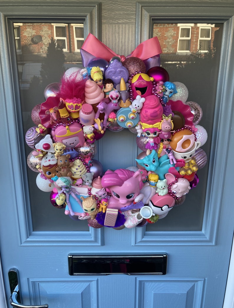 K&ouml;nnte beinhalten: Ein rosa-lilafarbener Weihnachtskranz, der mit verschiedenen kleinen Spielzeugen geschm&uuml;ckt ist, darunter ein Einhorn, ein rosa Pony, ein Donut und ein rosa Eiswaffel.