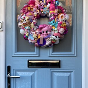 K&ouml;nnte beinhalten: Ein rosa-lilafarbener Kranz, der mit verschiedenen kleinen Spielzeugen geschm&uuml;ckt ist, darunter ein rosa Pony, ein blaues Einhorn und ein rosa-wei&szlig;es Eis am Stiel. Der Kranz h&auml;ngt an einer hellblauen T&uuml;r.