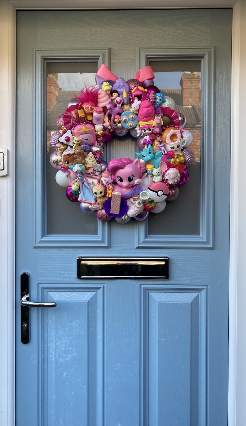 K&ouml;nnte beinhalten: Ein rosa-lilafarbener Kranz, der mit verschiedenen kleinen Spielzeugen geschm&uuml;ckt ist, darunter ein rosa Pony, ein blaues Einhorn und ein rosa-wei&szlig;es Eis am Stiel. Der Kranz h&auml;ngt an einer hellblauen T&uuml;r.