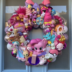 K&ouml;nnte beinhalten: Ein rosa-lilafarbener Weihnachtskranz, der mit verschiedenen Spielzeugen geschm&uuml;ckt ist, darunter ein rosa Pony, ein Einhorn, ein Drache und verschiedene Puppen. Der Kranz ist mit rosa und wei&szlig;en Kugeln und einer rosa Schleife verziert.
