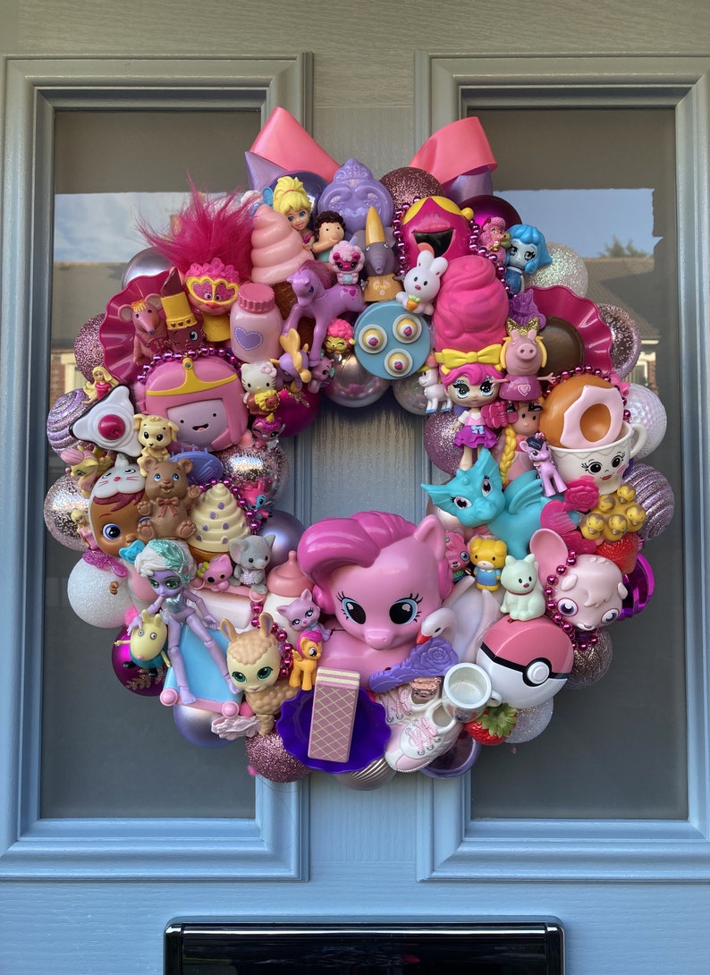 K&ouml;nnte beinhalten: Ein rosa-lilafarbener Weihnachtskranz, der mit verschiedenen Spielzeugen geschm&uuml;ckt ist, darunter ein rosa Pony, ein Einhorn, ein Drache und verschiedene Puppen. Der Kranz ist mit rosa und wei&szlig;en Kugeln und einer rosa Schleife verziert.