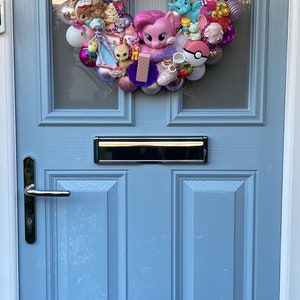 K&ouml;nnte beinhalten: Ein rosa-lilafarbener Kranz, der mit verschiedenen kleinen Spielzeugen und Schmuckst&uuml;cken verziert ist. Der Kranz besteht aus wei&szlig;em Schaumstoff und ist mit rosa-lilafarbenen B&auml;ndern geschm&uuml;ckt. Zu den Spielzeugen geh&ouml;ren ein Einhorn, ein Pony, eine Meerjungfrau, eine Prinzessin, eine Katze, ein Hund, ein Donut, ein Cupcake und eine Vielzahl anderer kleiner Spielzeuge.