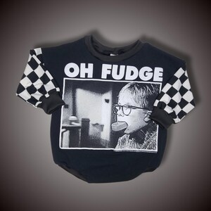 Może przedstawiać: Czarna bluza z szachownicą na rękawach. Z przodu bluzy znajduje się czarno-białe zdjęcie chłopca w okularach trzymającego w ustach kawałek mydła. Nad zdjęciem nadrukowany jest tekst "OH FUDGE".
