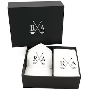 May include: A black gift box with a white golf hat and towel inside. The hat and towel both have the letters "R" and "A" with crossed golf clubs.