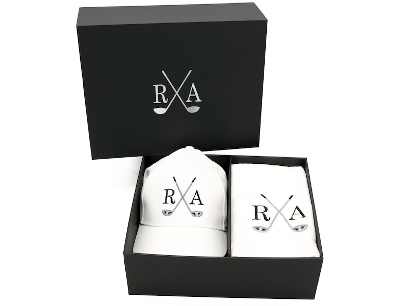 May include: A black gift box with a white golf hat and towel inside. The hat and towel both have the letters "R" and "A" with crossed golf clubs.