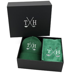 May include: A black gift box with a green golf hat and towel inside. The hat and towel both have the initials "TH" and a pair of crossed golf clubs embroidered on them.