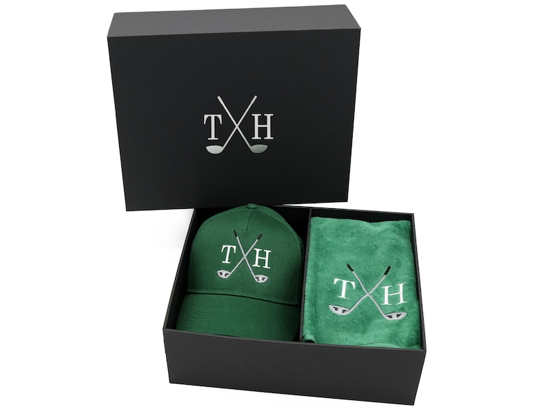 May include: A black gift box with a green golf hat and towel inside. The hat and towel both have the initials "TH" and a pair of crossed golf clubs embroidered on them.