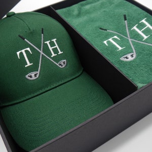 May include: A green golf hat and towel set with the initials "TH" and crossed golf clubs embroidered on both items. The set is packaged in a black box.
