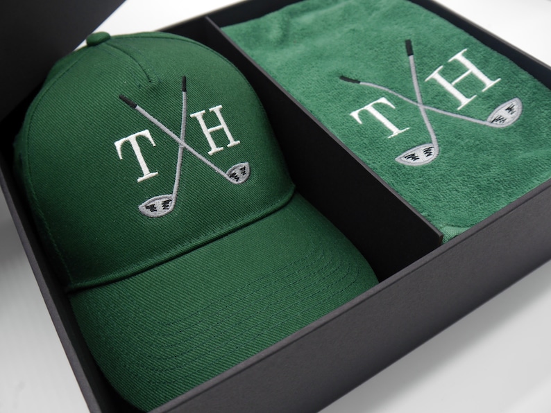 May include: A green golf hat and towel set with the initials "TH" and crossed golf clubs embroidered on both items. The set is packaged in a black box.