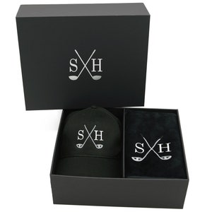 May include: A black gift box with a black golf cap and a black golf towel inside. The box, cap, and towel all have the same silver embroidered logo: crossed golf clubs with the letters "SH".