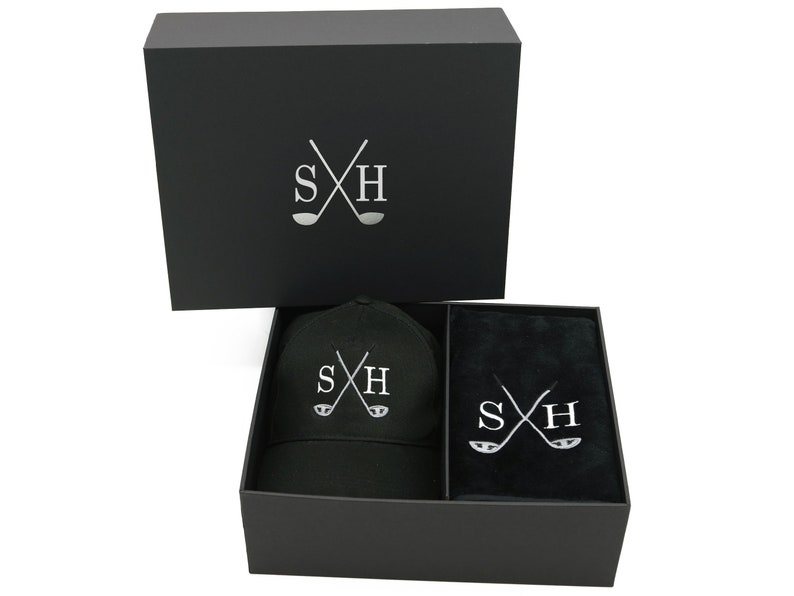 May include: A black gift box with a black golf cap and a black golf towel inside. The box, cap, and towel all have the same silver embroidered logo: crossed golf clubs with the letters "SH".