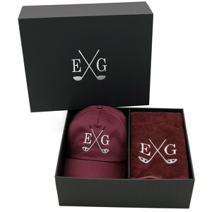 May include: A black gift box with a burgundy golf cap and towel. The box has the letters "EXG" with crossed golf clubs on the lid. The cap and towel also have the same logo embroidered on them.