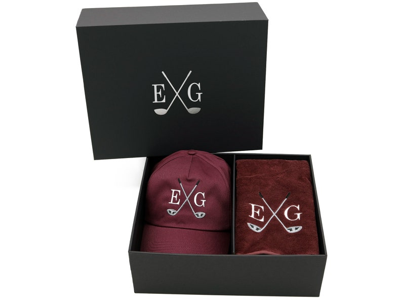 May include: A black gift box with a burgundy golf cap and towel. The box has the letters "EXG" with crossed golf clubs on the lid. The cap and towel also have the same logo embroidered on them.