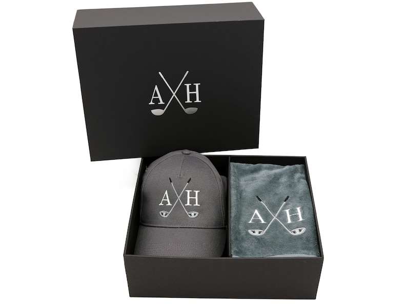 May include: A black gift box with a gray baseball cap and a gray towel inside. The cap and towel both have the letters "AXH" embroidered on them with crossed golf clubs.