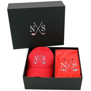 May include: A black gift box with a red golf hat and a red towel inside. The hat and towel both have the letters "NS" and a pair of crossed golf clubs embroidered on them.