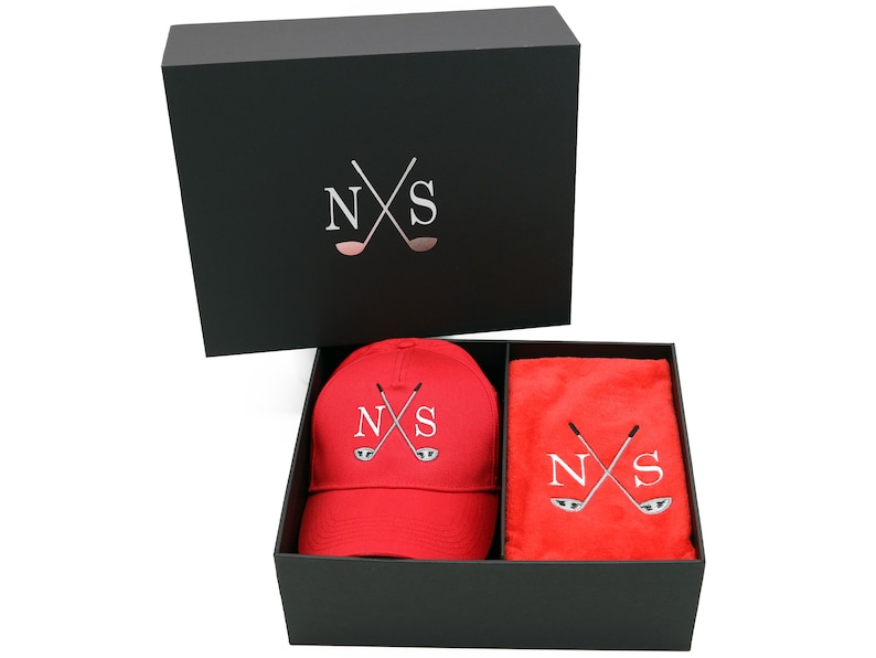 May include: A black gift box with a red golf hat and a red towel inside. The hat and towel both have the letters "NS" and a pair of crossed golf clubs embroidered on them.