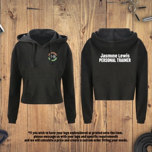 May include: Black cropped hoodie with a white logo that says "Jasmine Lewis PERSONAL TRAINER" on the back and a green, white, and black logo on the front. The hoodie is on a wooden background.
