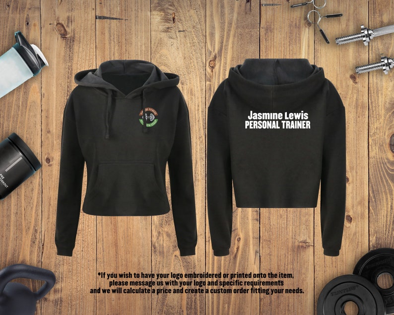 May include: Black cropped hoodie with a white logo that says "Jasmine Lewis PERSONAL TRAINER" on the back and a green, white, and black logo on the front. The hoodie is on a wooden background.