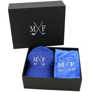 May include: A black gift box with a blue golf hat and a blue towel inside. The hat and towel both have the initials "MF" and crossed golf clubs embroidered on them.
