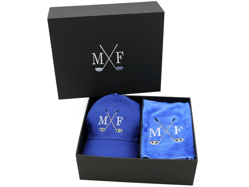 May include: A black gift box with a blue golf hat and a blue towel inside. The hat and towel both have the initials "MF" and crossed golf clubs embroidered on them.