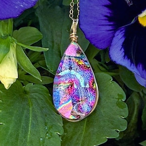 May include: A teardrop-shaped pendant necklace with a gold-colored chain. The pendant features a colorful, iridescent design with shades of pink, blue, and purple. The pendant is set against a backdrop of green leaves and purple flowers.