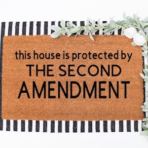 May include: A brown coir doormat with black text that reads "this house is protected by THE SECOND AMENDMENT". The doormat is on a black and white striped rug.