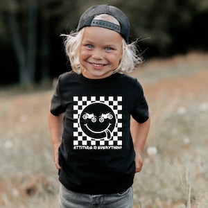 May include: Black t-shirt with a white checkered pattern and a smiley face with dirt bike wheels for eyes. The text "ATTITUDE IS EVERYTHING" is printed below the smiley face.