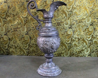 May include: An antique silver-coloured pitcher with an elaborate handle and spout. The body of the pitcher is decorated with relief patterns and stands on a pedestal base. The background is a gold and olive paisley pattern.