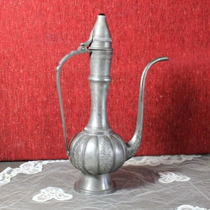 May include: A silver pitcher with an ornate design and a hinged lid. The pitcher has a long spout and a round body. It is sitting on a white lace tablecloth.