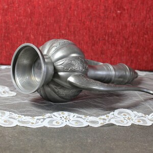 May include: A silver metal pitcher with an ornate design and a long spout. The pitcher is lying on a white lace tablecloth.