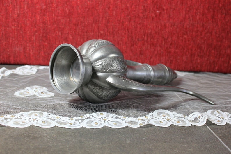 May include: A silver metal pitcher with an ornate design and a long spout. The pitcher is lying on a white lace tablecloth.