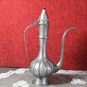 May include: A silver teapot with an ornate design and a long spout. The teapot is sitting on a white lace tablecloth.