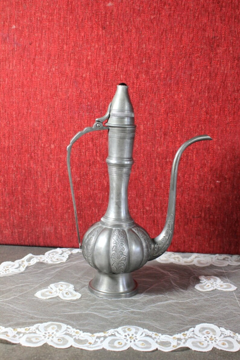 May include: A silver teapot with an ornate design and a long spout. The teapot is sitting on a white lace tablecloth.